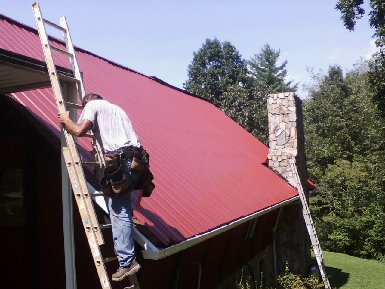 Roofer on a ladder beside a red metal roof and stone chimney — Soffit & Fascia Repair by Roofing Brothers in Seven Valleys
